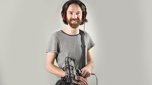 A man with a beard wearing headphones and a striped t-shirt, holding audio equipment.