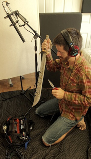 A man in a plaid shirt and headphones holds a sword and a knife near a microphone setup.