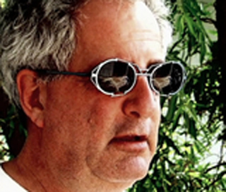 Close-up of a man with gray hair wearing round sunglasses that reflect birds in the lenses.