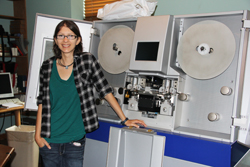 A woman in a plaid shirt and green top stands beside a large, open piece of scientific machinery.