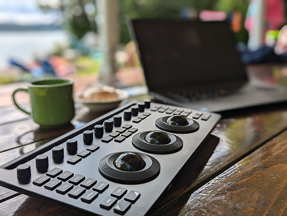 A black video editing control surface with knobs and trackballs sits on a wooden table near a green mug.