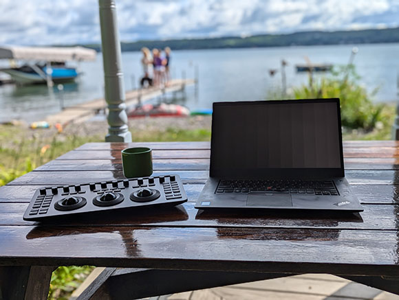 A black laptop and a control surface sit on a wet wooden table with a lake and people in the background.