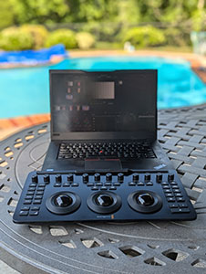 A black laptop and a music production controller sit on a woven outdoor table near a swimming pool.