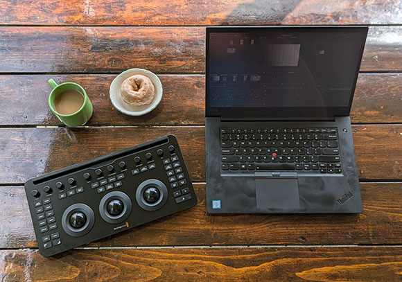 A black laptop, a black control surface with dials, a green mug of coffee, and a donut on a wooden table.