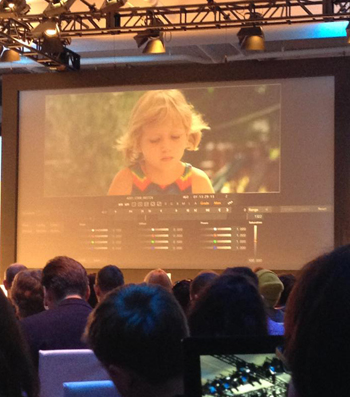 A large projection screen shows a close-up of a blonde child, viewed from the back of a darkened audience.
