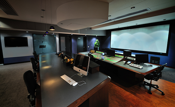 A dark media production studio featuring a large projection screen, mixing consoles, and computer workstations.
