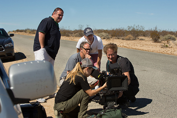 A film crew operates a professional camera on a tripod by a desert road under a clear blue sky.