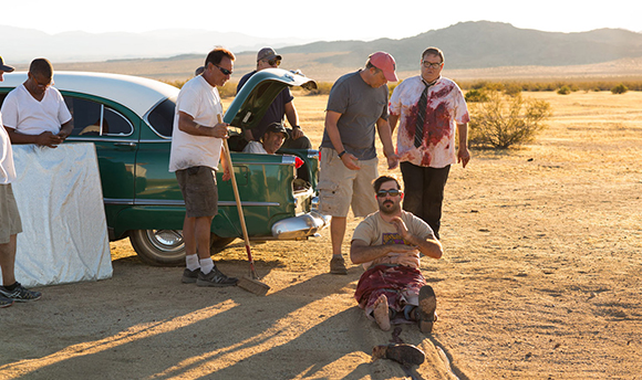 Men stand near a green vintage car in a desert landscape during sunset.