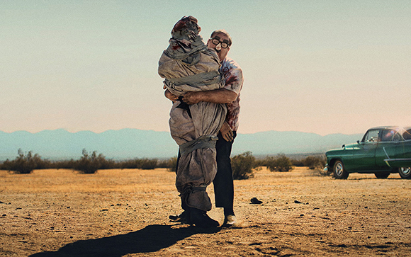 A man in glasses embraces a large, blood-stained figure wrapped in cloth in a desert setting.