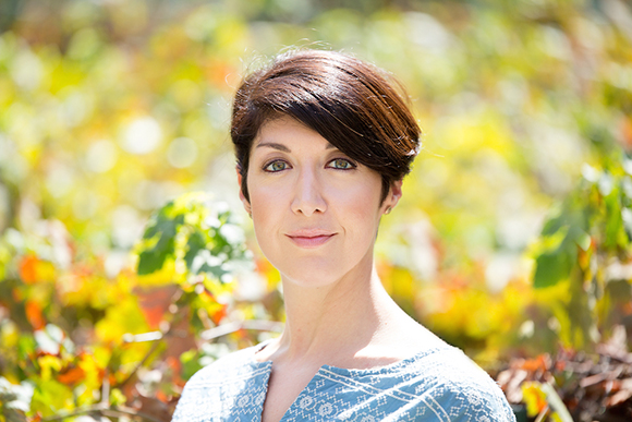 A woman with short dark hair smiling in front of a blurred background of green and yellow foliage.