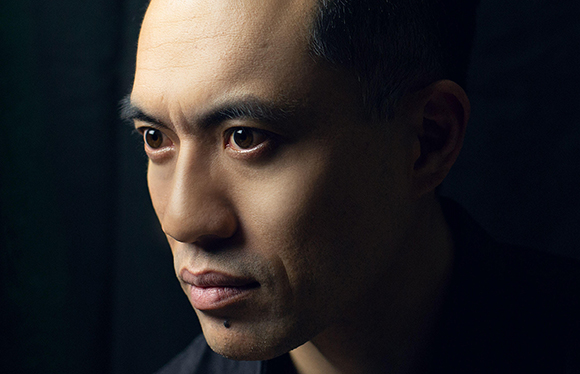 Close-up portrait of a man with dark hair and brown eyes looking toward the left against a dark background.