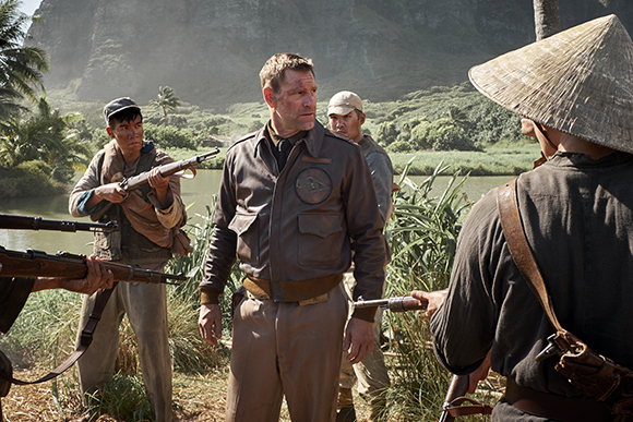 Men in mid-20th century military and civilian attire stand near a river with rifles in a tropical setting.