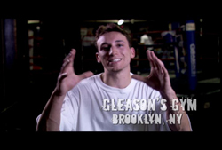 A smiling man in a white t-shirt gestures with his hands in a dark boxing gym.