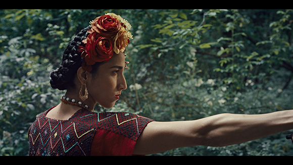 A woman with braided hair adorned with red and yellow flowers looks sideways against a leafy green background.