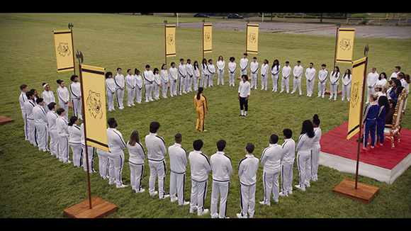 A large group of people in white tracksuits stand in a circle on a grass field around a person in yellow.