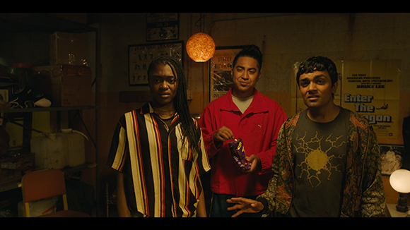 Three young people stand together in a cluttered, dimly lit room with posters on the wall.