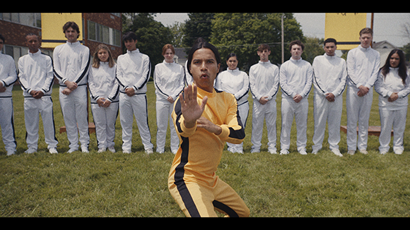 A person in a yellow tracksuit performs a martial arts pose in front of a line of people in white tracksuits.