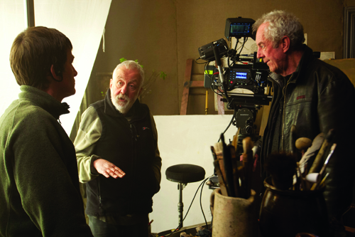 Three men stand in a studio near a large professional video camera and art supplies.