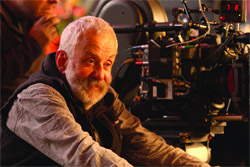 An older man with a grey beard looking intently at a large professional motion picture camera.