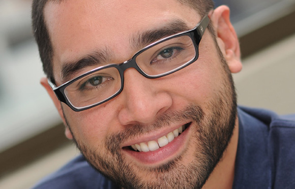 Close-up of a smiling man with dark hair, a beard, and black-rimmed glasses.