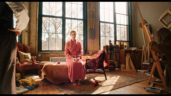 A woman in a pink robe sits on a stool in a sunlit art studio filled with easels and canvases.