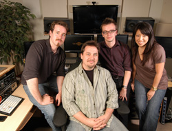 Four people smiling and posing in a studio filled with monitors and audio equipment.