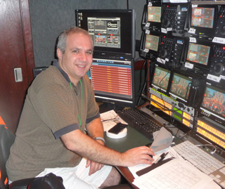 A man smiling while sitting in front of multiple computer monitors and broadcast equipment.