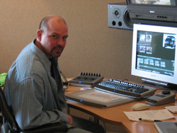 A man in a striped shirt sits at a desk with a computer monitor, keyboard, and audio mixer.