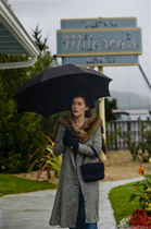 A woman in a grey coat and fur collar holds a black umbrella outdoors near a restaurant sign.