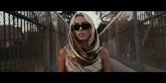 A blonde woman wearing dark sunglasses and a draped gold metallic hood stands between two metal fences.
