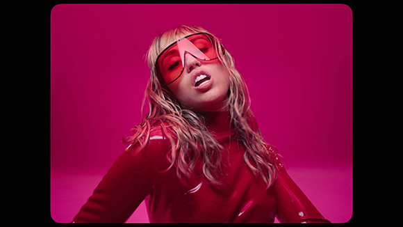 A person in a red turtleneck and large red sunglasses poses against a solid pink background.