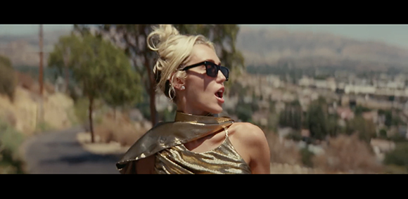 A woman with blonde hair in a bun wearing dark sunglasses and a metallic gold top outdoors.