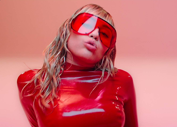 A person wearing a shiny red latex top and oversized red-tinted sunglasses against a pink background.