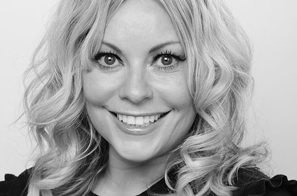 A smiling woman with wavy blonde hair and dark eyeliner looking toward the camera.
