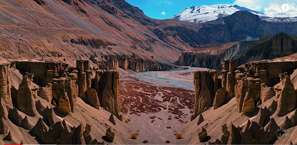 Jagged rock pillars frame a wide, reddish riverbed leading toward distant mountains and a snow-capped peak.