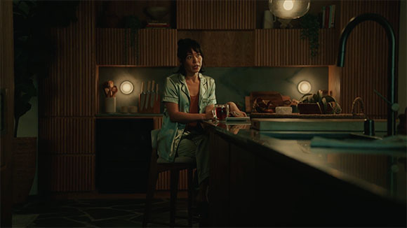 A woman sits at a dark kitchen island holding a glass, looking upward in a dimly lit room.
