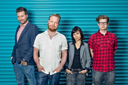 Four people standing in a row against a bright blue corrugated metal background.