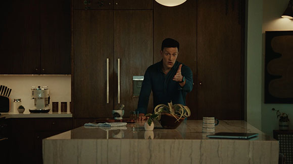 A man in a dark blue shirt stands behind a kitchen island, pointing his finger forward with a serious expression.