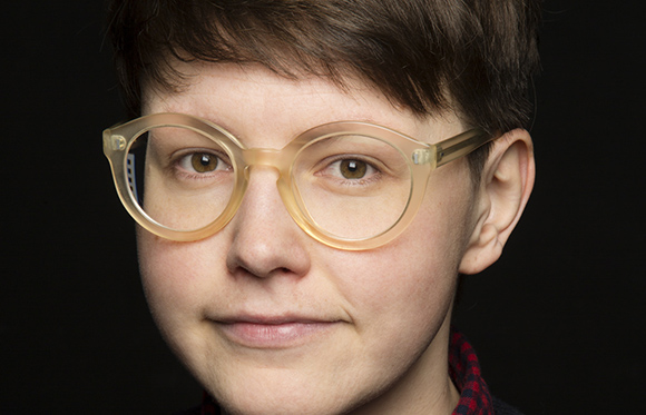 A person with short brown hair and thick, translucent yellow glasses looking forward against a black background.