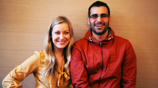 A smiling man in glasses and a red jacket standing next to a smiling woman in a tan shirt.