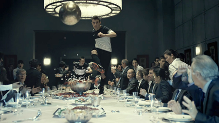 A young man in athletic wear kicks a soccer ball over a long, crowded dinner table in a formal dining room.
