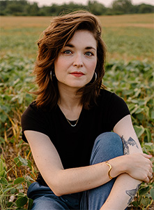 A woman with brown hair wearing a black t-shirt and blue jeans sits in a green field.