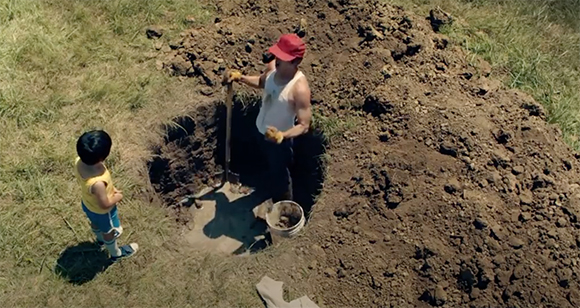 A man in a red cap digs a hole in a grassy field while a young child in a yellow shirt watches from the side.