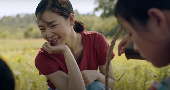 A smiling woman in a red shirt rests her chin on her hand while looking at a child in a field.