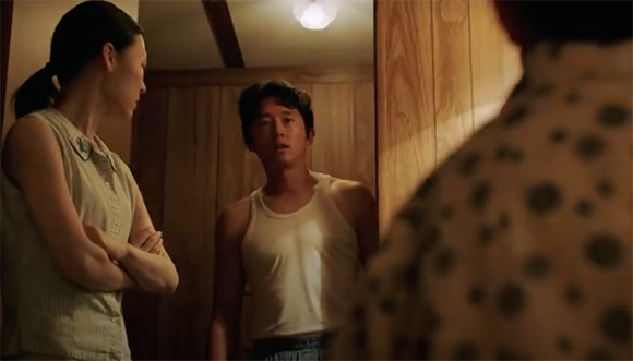 A man in a white tank top looks toward a woman with crossed arms in a wood-paneled room.