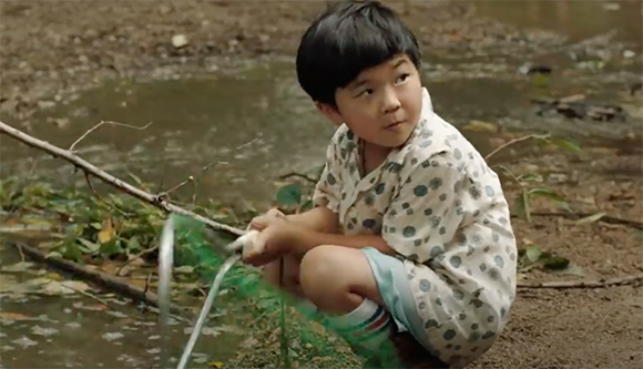A young child in a patterned shirt squats by a muddy puddle holding a green net.