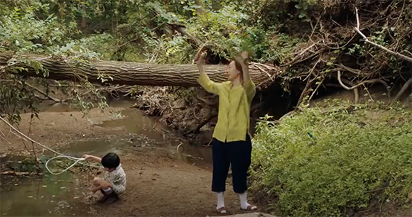 A woman in a yellow shirt stands by a fallen log while a child crouches near a stream in a forest.
