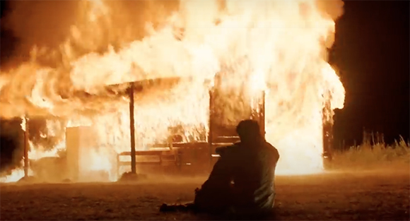 A silhouette of a person sitting on the ground facing a large building engulfed in intense orange flames.