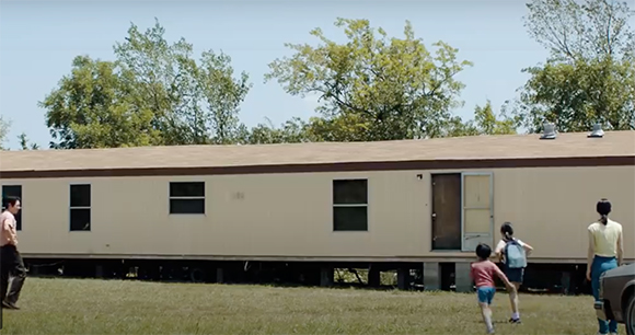 A tan mobile home sits on a grassy field with several people walking nearby under a clear sky.