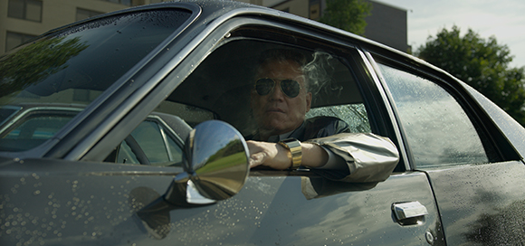 A man wearing sunglasses drives a dark car through a light rain.
